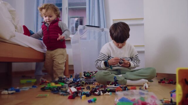 Young child constructs a toy creation from numerous parts across the room as his toddler brother steps forward with growing stability, showing focus and physical confidence