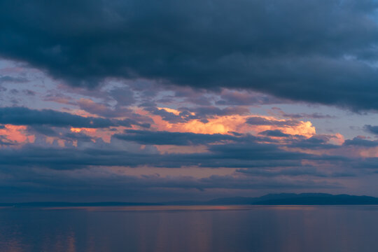 Dramatic Adriatic seascape at dusk with calm water, distant mountainous islands and layered storm clouds illuminated by soft pink, orange and purple evening light