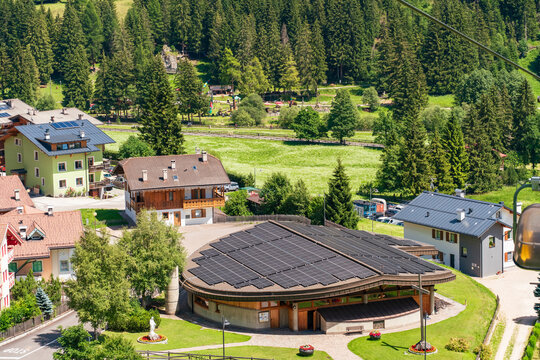 Elevated view over Pera di Fassa, showing traditional alpine houses, green meadows and dense conifer forests, with a large modern building equipped with extensive rooftop solar panels