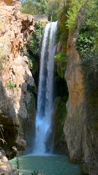 Plano general vertical de una alta cascada cayendo desde un ca&ntilde;&oacute;n rocoso hacia una poza de agua turquesa en un entorno natural soleado con vegetaci&oacute;n. Zaragoza.