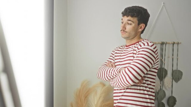 Man in red striped pyjama shirt with arms crossed, standing by macrame wall hanging and pampas grass in building; quiet contemplation.