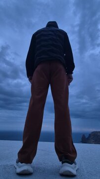 Young man stands overlooking a vast ocean cliffside at twilight. Low angle tracking shot.