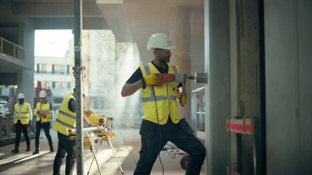 Real Estate Building Construction Site: Skilled Specialist Drills Wall Using Diamond Core Machine While Colleagues Cut Timber and Review Plans Behind Him on Apartment Project. Wide Tracking Shot