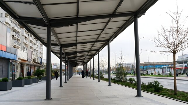 Modern covered walkway with metal supports and tiled pavement  lined with planters and bare trees under overcast sky