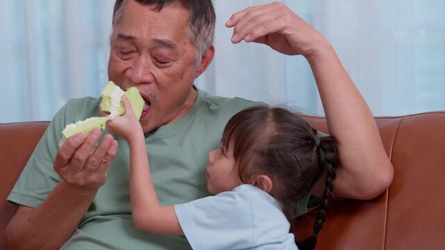 Grandfather and granddaughter sharing a piece of fruit together at home. The intimate family moment conveys affection, caregiving, healthy eating, childhood, and intergenerational bonding.