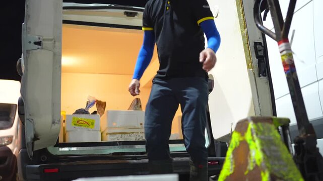 Worker unloading fresh fish crates from refrigerated van at wholesale seafood market during night shift.