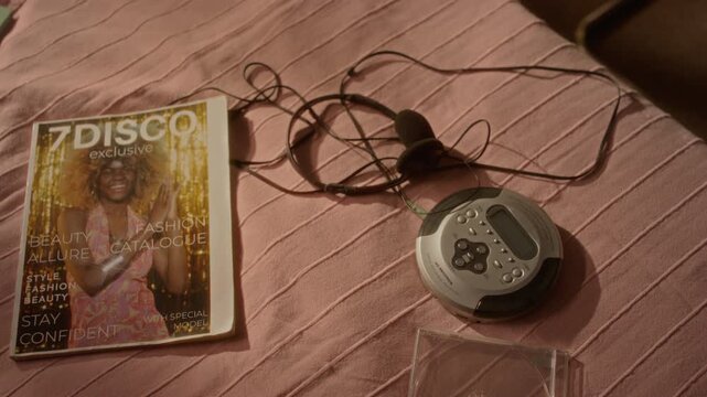 Retro portable CD player with headphones and fashion magazine lying on pink bedsheet, top view. Ideal for nostalgia marketing, retro music documentaries, or vintage lifestyle commercials