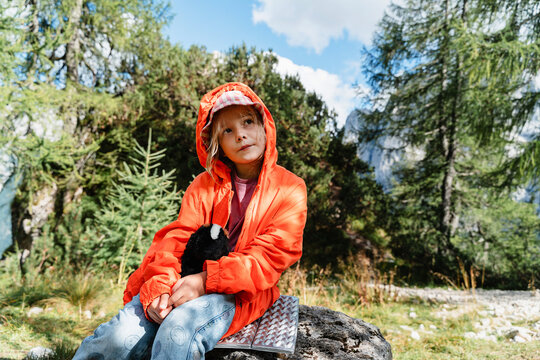 Cute girl in orange hoodie resting on rock in sunny pine forest. Happy family on summer holiday and active outdoor lifestyle.
