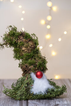Handmade Christmas gnome made of natural moss with a red nose on a wooden background and soft bokeh lights.