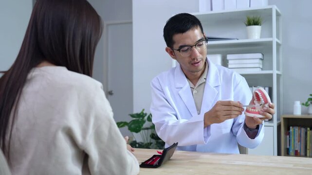 Asian male dentist holding a teeth model explaining oral care to a female doctor during a medical podcast interview. Recording a dental healthcare audio broadcast in a modern clinic studio.