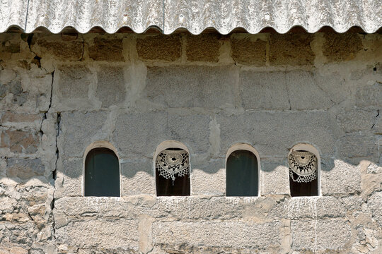 Vusanje, Gusinje Municipality, Montenegro - 28 July 2005: View of a weathered stone wall with four small arched windows, some featuring delicate lace curtains, under a corrugated roof.