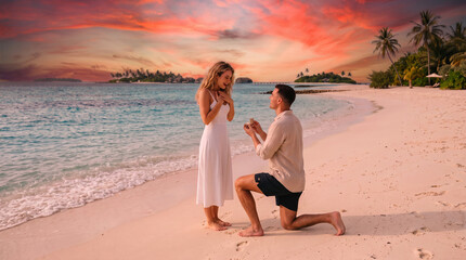 Fototapeta premium Romantic marriage proposal on a pristine Maldives beach: a young man kneeling on the white sand to propose to his surprised girlfriend with a ring.