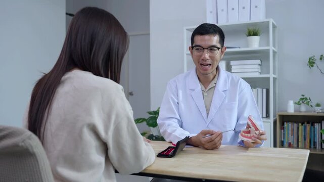 Asian male dentist holding a teeth model explaining oral care to a female doctor during a medical podcast interview. Recording a dental healthcare audio broadcast in a modern clinic studio.