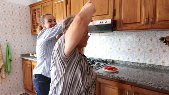 Joyful elderly mother and her adult daughter dancing and laughing together in a cozy home kitchen, bonding and having fun while preparing a meal, expressing love and happiness