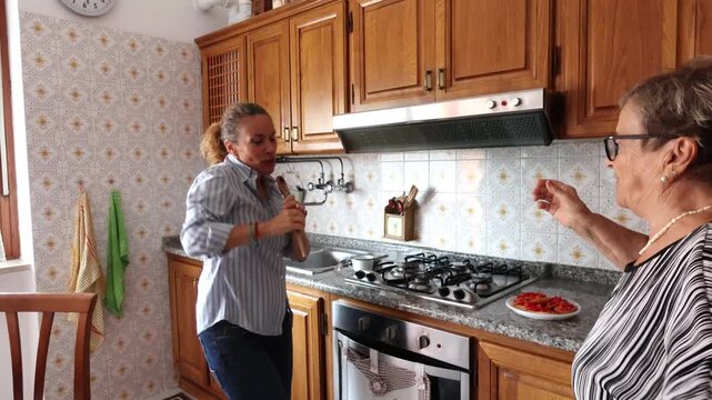 Playful adult daughter singing with a wooden spoon and dancing with her elderly mother while preparing a meal together, sharing a happy and loving moment of intergenerational bonding at home