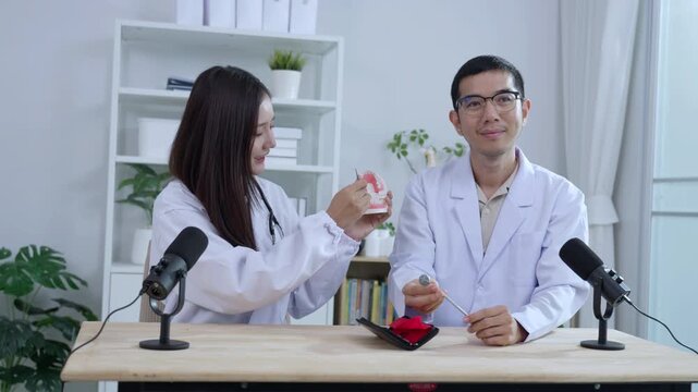 Asian male dentist holding a teeth model explaining oral care to a female doctor during a medical podcast interview. Recording a dental healthcare audio broadcast in a modern clinic studio.