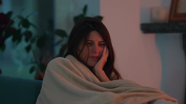 Tired woman sits wrapped in a soft blanket on the living room sofa with her cheek resting on one hand, expressing fatigue, contemplation and a need for comfort