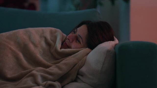 Resting woman reclines beneath a cozy blanket with her head supported by a pillow on the living room couch, embracing peaceful relaxation and physical renewal