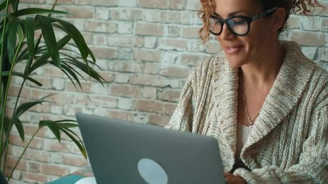 Concentrated middle-aged businesswoman wearing glasses and working on her laptop from home, enjoying a modern remote job and smart working lifestyle with a brick wall in the background