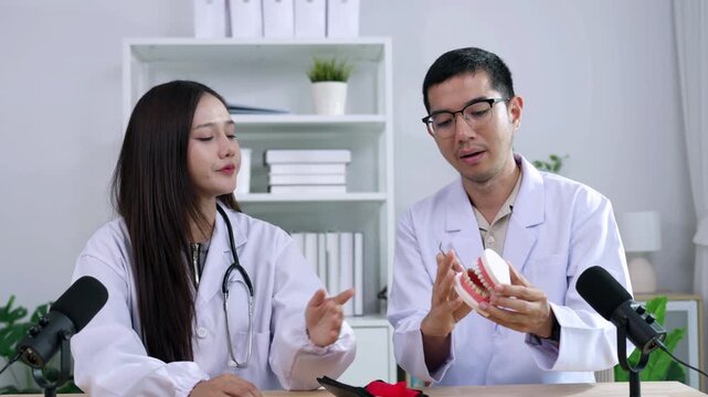 Asian male dentist holding a teeth model explaining oral care to a female doctor during a medical podcast interview. Recording a dental healthcare audio broadcast in a modern clinic studio.