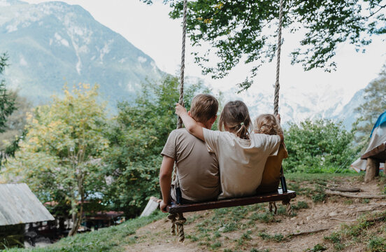 Three joyful children sitting together on a wooden tree swing overlooking a mountain valley.
