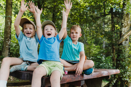 Three joyful children sitting on a picnic table raising their arms in celebration at a forest camp.