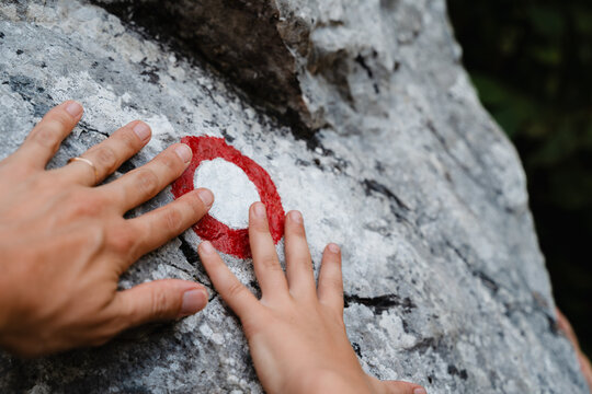 Close-up of adult and child hands touching a red and white painted trail marker on a large rock.