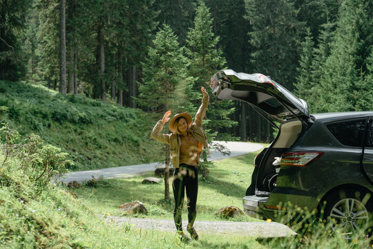 Happy young woman in a hat closing car trunk on a forest road during a joyful summer road trip adventure.