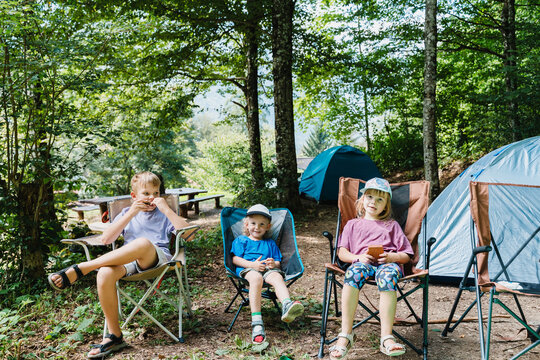 Three kids relaxing in camping chairs next to tents in a sunny green forest.