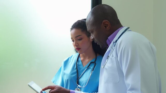 A male doctor and a female nurse reviewing patient information together on a digital tablet near a window.