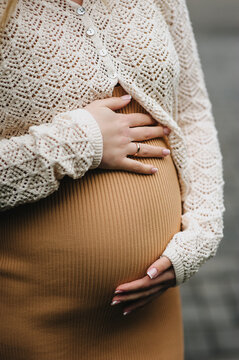 A close-up photo of a young pregnant woman in a dress, gently holding her large belly in her hands. Waiting for a miracle. Pregnancy concept.