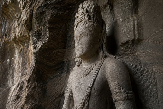 An ancient unfinished stone statue of a deity with a worn face in a rocky alcove.