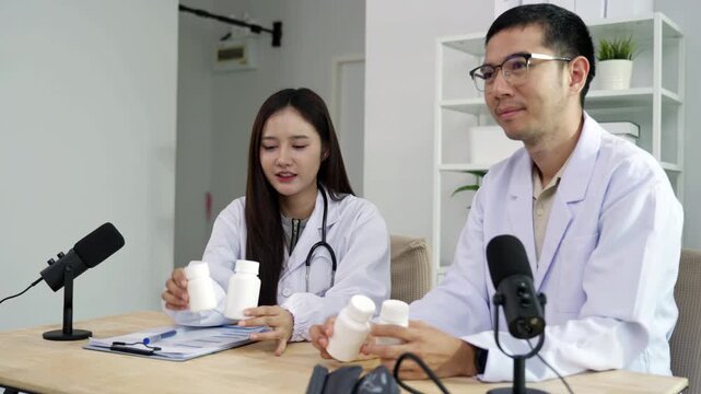 Asian male doctor explaining medicine from a blank white bottle to a female doctor during a medical podcast interview. Recording a healthcare audio broadcast in a clinic. Pharmacy concept.