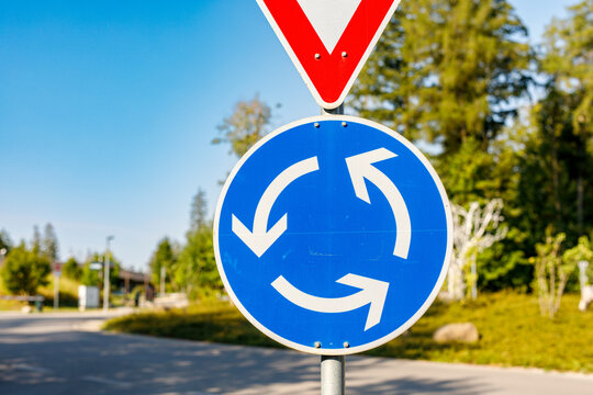 Blue circular roundabout sign with yield sign above, set against a clear blue sky and green trees.