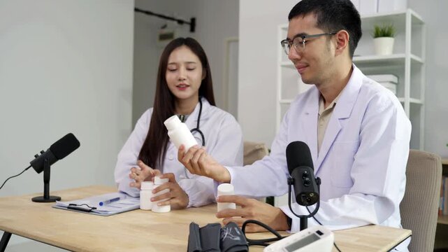 Asian male doctor explaining medicine from a blank white bottle to a female doctor during a medical podcast interview. Recording a healthcare audio broadcast in a clinic. Pharmacy concept.
