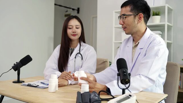 Asian male doctor explaining medicine from a blank white bottle to a female doctor during a medical podcast interview. Recording a healthcare audio broadcast in a clinic. Pharmacy concept.