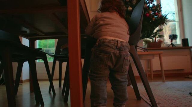 Toddler stepping down from dining chair beside decorated Christmas tree in family home, carefully lowering one leg toward the floor during an indoor holiday moment