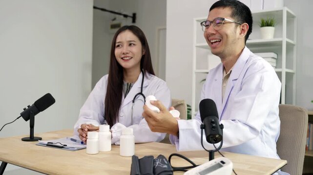 Asian male doctor explaining medicine from a blank white bottle to a female doctor during a medical podcast interview. Recording a healthcare audio broadcast in a clinic. Pharmacy concept.