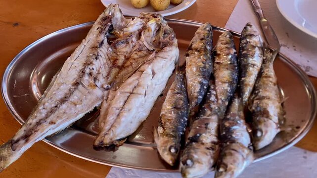 Plate of grilled fish with sardines and mackerel served on metal tray on wooden table.