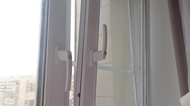 The hand closes the window with a key. Closing a white PVC window. Double handles, glass panels, light condensation on the frame.