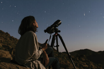 Fototapeta premium A woman observes a comet through a telescope on a rocky landscape, highlighting the intersection of nature and human curiosity during a breathtaking cosmic event.