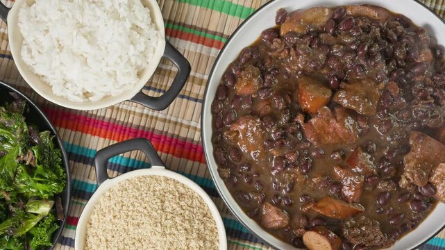 Zoom in shot of feijoada on a table surrounded by side dishes, kale, farofa and rice.