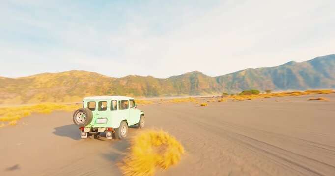 FPV aerial view of a light blue off-road vehicle driving across the vast sandy volcanic plain of Mount Bromo in Indonesia.