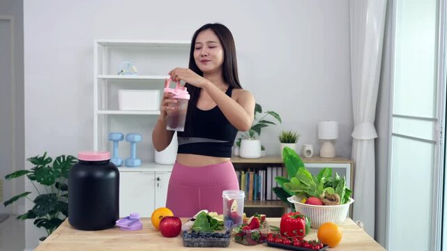 Smiling Asian fitness influencer recording a health podcast, scooping pink protein powder into a shaker bottle. Woman vlogger creating sports nutrition and healthy diet content with fresh food.