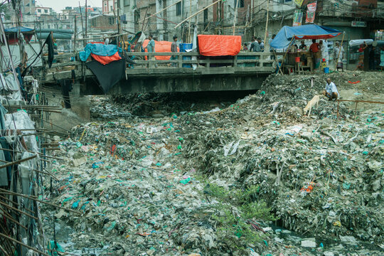 Dhaka, Bangladesh - 15 June 2026: View of a heavily polluted canal filled with plastic waste and debris near the Buriganga River, with a bridge and makeshift structures in the background.