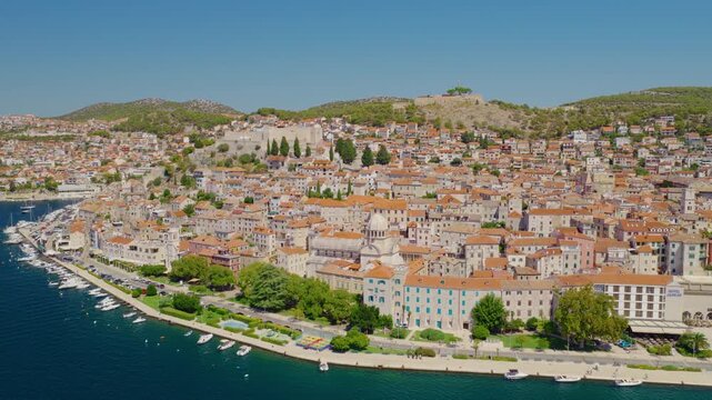 Aerial drone view of &Scaron;ibenik, Croatia, with a smooth backward camera movement revealing the historic waterfront, dense red rooftops, medieval architecture, blue Adriatic Sea, boats and hills.