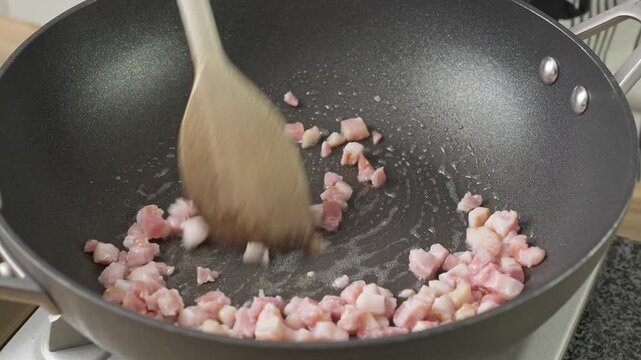 Cooking bacon/pancetta lardons in a non-stick wok. Real-time food preparation footage showing diced pancetta sizzling, rendering fat, and browning over medium heat in a kitchen pan.