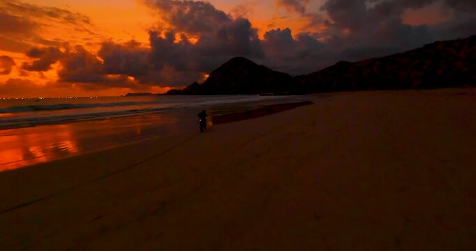 FPV aerial view of a sandy beach at sunset with dramatic orange clouds over the ocean and distant mountains in Bali, Indonesia.