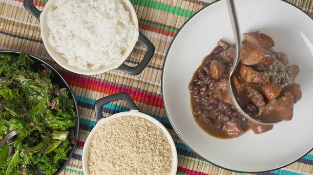 Zoom in shot of feijoada being served in a bowl on a table surrounded by side dishes, kale, farofa and rice.