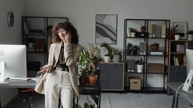 Young adult Caucasian businesswoman talking on smartphone in modern office, gesturing and smiling. Video is suitable for corporate communication and remote work marketing campaigns